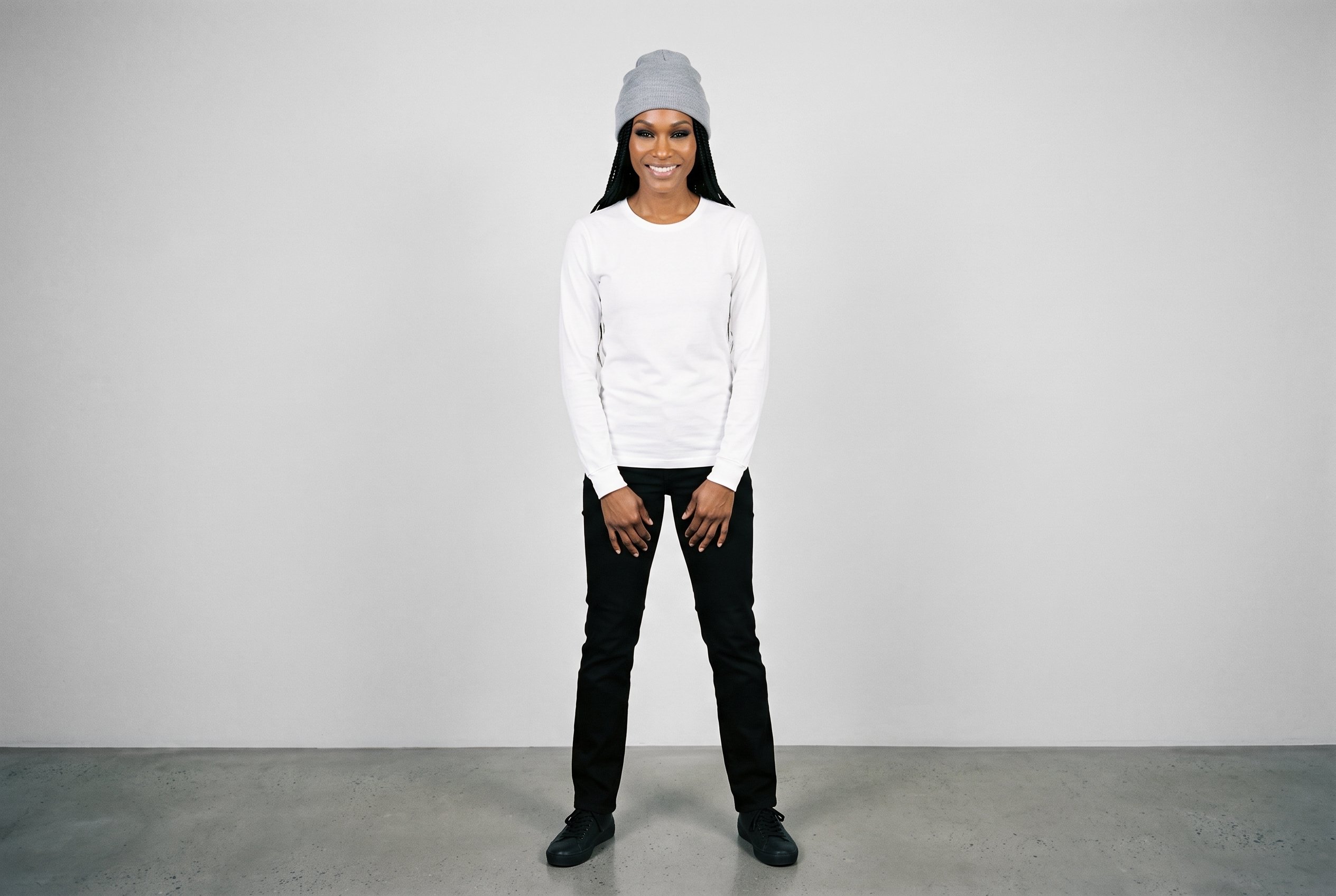 A 42-year-old African woman wears a women's grey ribbed beanie in a full-body front view. She is smiling with her hands held in front, against a fog grey studio-wall backdrop.