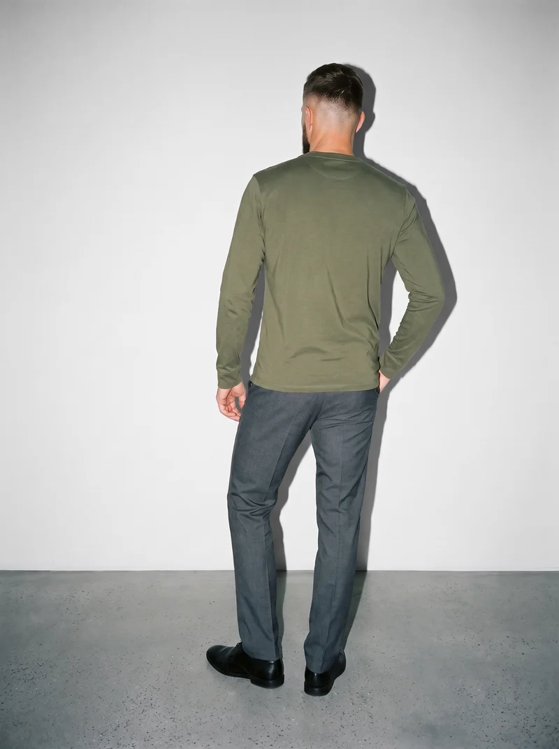 A 37-year-old Slavic man wears an olive long-sleeve Henley top in a full-body back view, with his weight positioned back. A fog-grey studio wall forms the backdrop.
