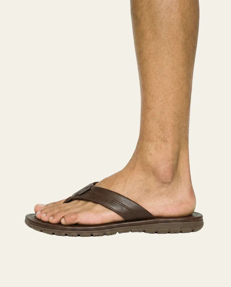 Men's brown flip-flops shown from the side with the toe pointed, framed at the ankle against an alabaster flat-color backdrop.