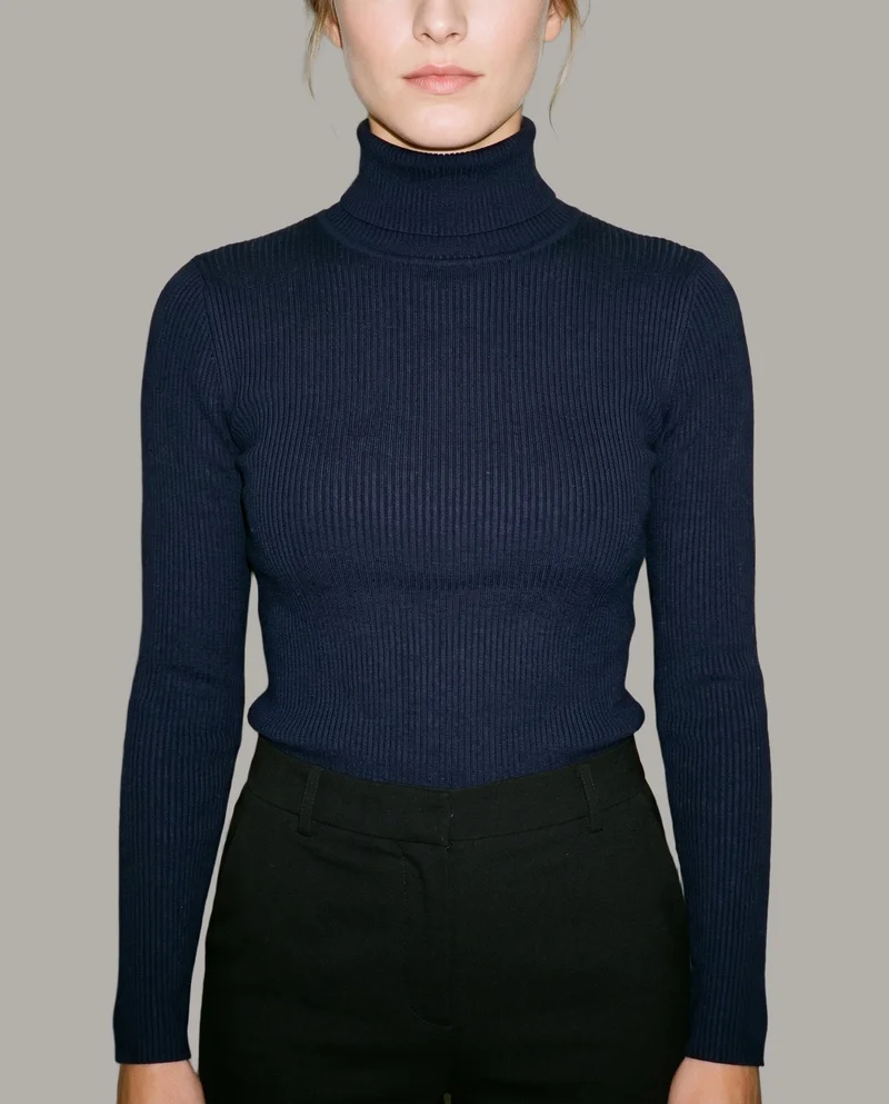 A woman models a women's navy ribbed turtleneck in a front-facing torso crop with her arms relaxed. The garment is shown against a flat Couture Grey backdrop.