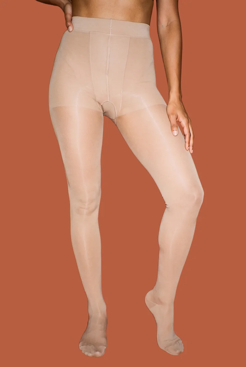 A female model’s lower body is shown from the front wearing nude sheer tights, with her knees positioned inward. A flat terracotta clay backdrop fills the background.