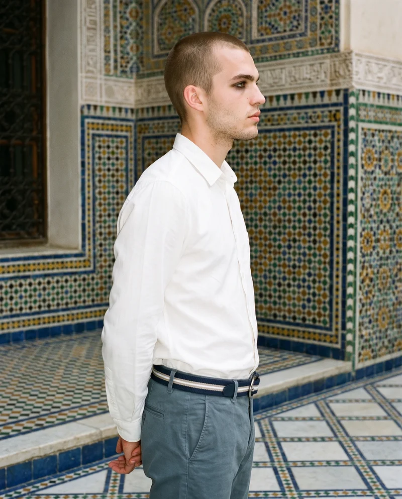 A smiling 20-year-old Caucasian man is shown in a three-quarter-length side view with his hands behind his back, wearing a men's striped woven belt. A Moroccan riad courtyard serves as the backdrop.