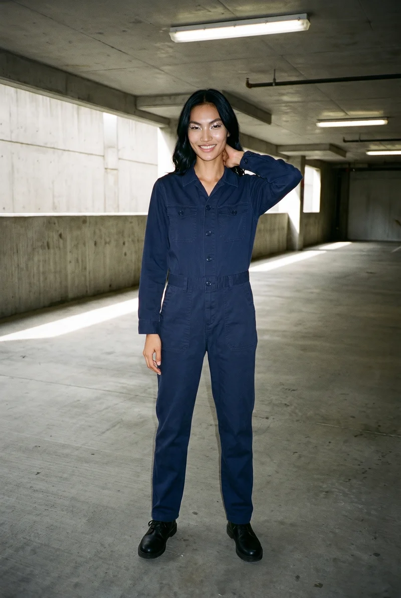 A 20-year-old Southeast Asian woman wears a navy cotton boilersuit in a front-facing environmental view. She is smiling with one hand behind her neck, with a concrete parking garage as the backdrop.