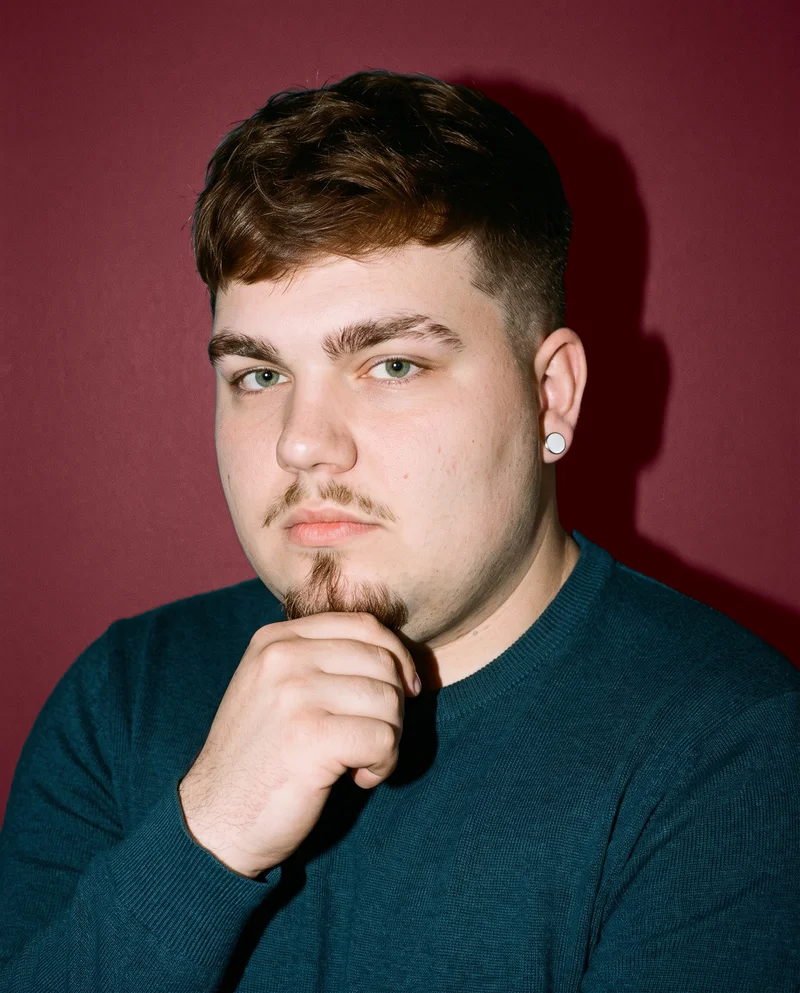 A 21-year-old Caucasian man is shown in a bust-length front three-quarter view with a neutral expression and one hand at his jaw. He is wearing men's silver stud earrings against a wine-red studio wall backdrop.
