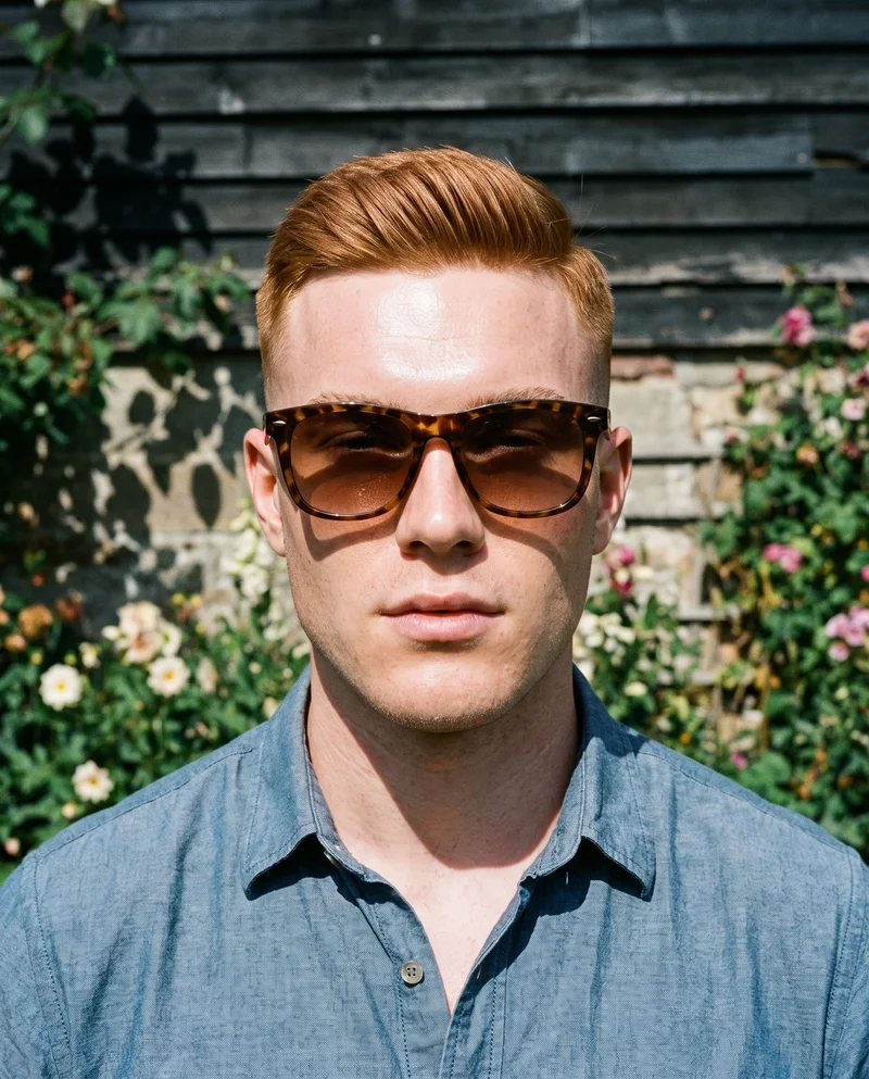 A 23-year-old Caucasian man is shown from the bust in a front view with his shoulders square and a soft facial expression, wearing men's tortoiseshell Wayfarer sunglasses. A Cottage Garden serves as the backdrop.