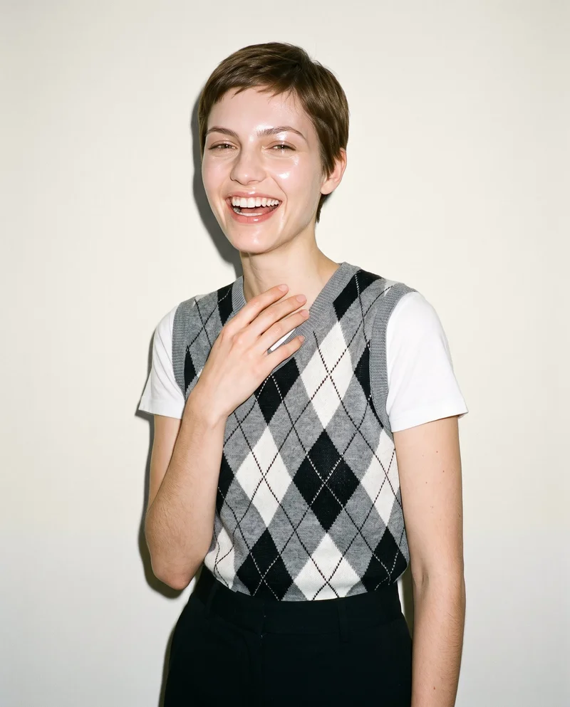 A 22-year-old Caucasian woman is shown from the upper body at an angled front view, wearing a grey argyle sweater vest with one hand at her collarbone. She is laughing against an alabaster studio-wall backdrop.
