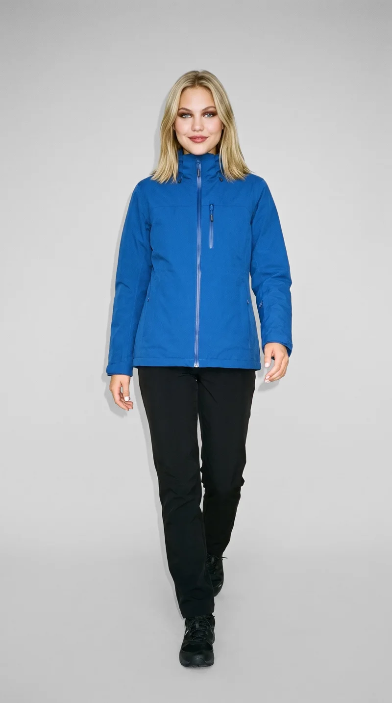 A 20-year-old Caucasian woman is shown full body from the front, smiling as she walks while wearing a blue waterproof ski jacket. The image uses a flat pearl grey backdrop.