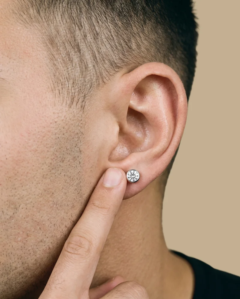 A crystal stud earring is shown on a man's ear in side view, with a finger touching the earlobe. The ear and earring are framed against a flat khaki backdrop.