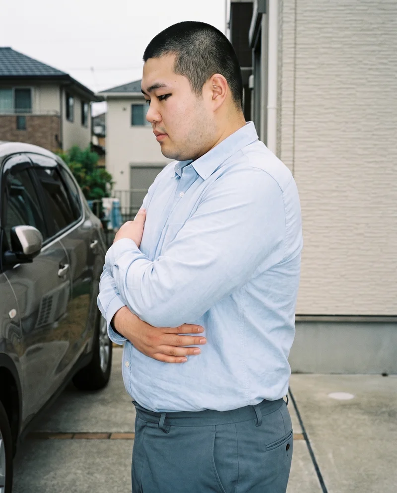 A 22-year-old Japanese man wears a light blue Oxford shirt in an upper-body side view. He stands in a self-hug pose with a neutral expression, against a suburban driveway backdrop.