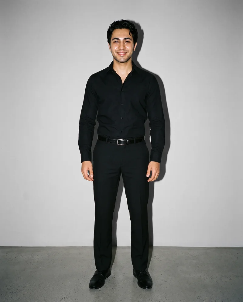 A 22-year-old Arab man is shown in an environmental front view, smiling and standing with his weight shifted back while wearing a men's reversible leather belt. A dove grey studio-wall backdrop frames the scene.