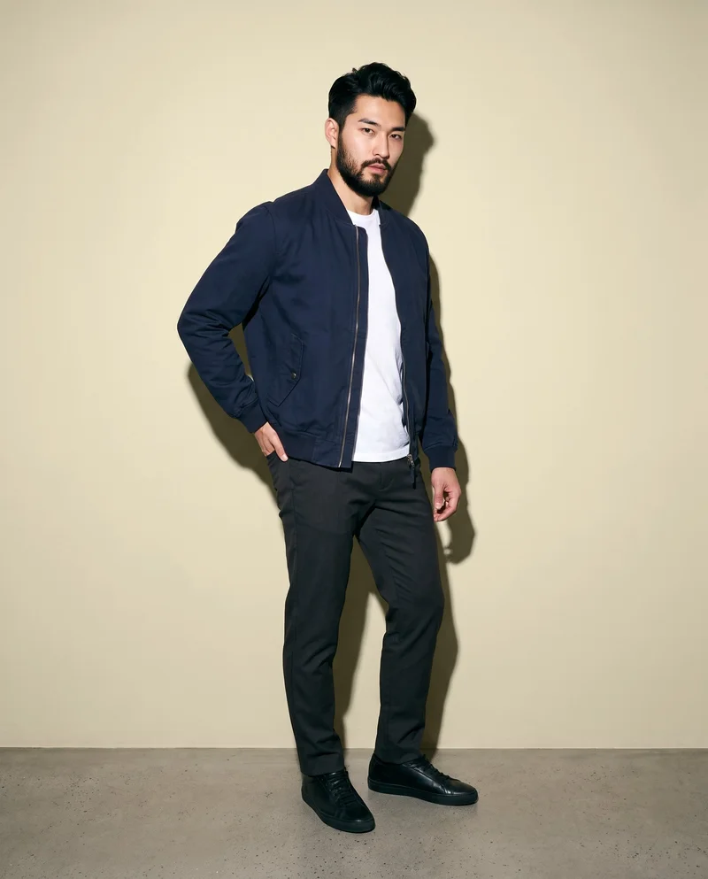 A 25-year-old Japanese man wears a men's navy bomber jacket in a full-body, angled-front view. He stands with one hand on his hip and a serious expression against a wheat-field backdrop.