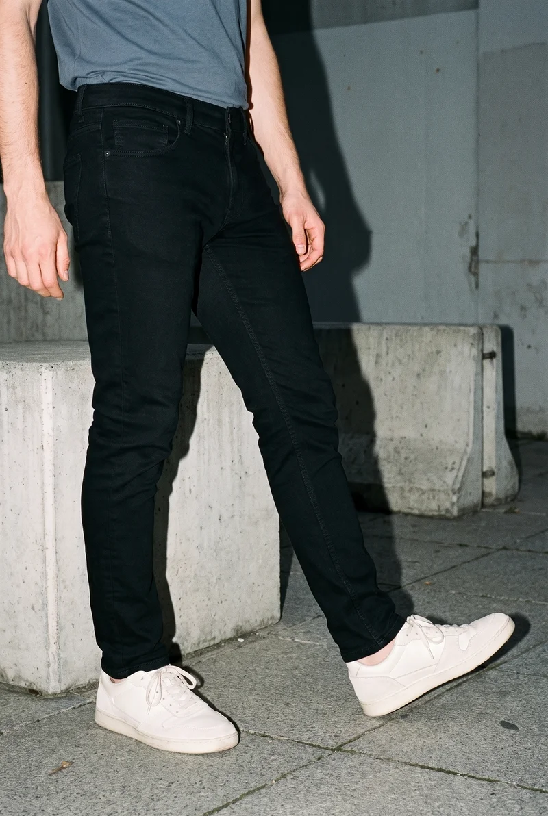 A male model wears black skinny jeans in a lower-body view, posed with one foot forward and shown from a front three-quarter angle. Parkour-style urban obstacles form the backdrop.