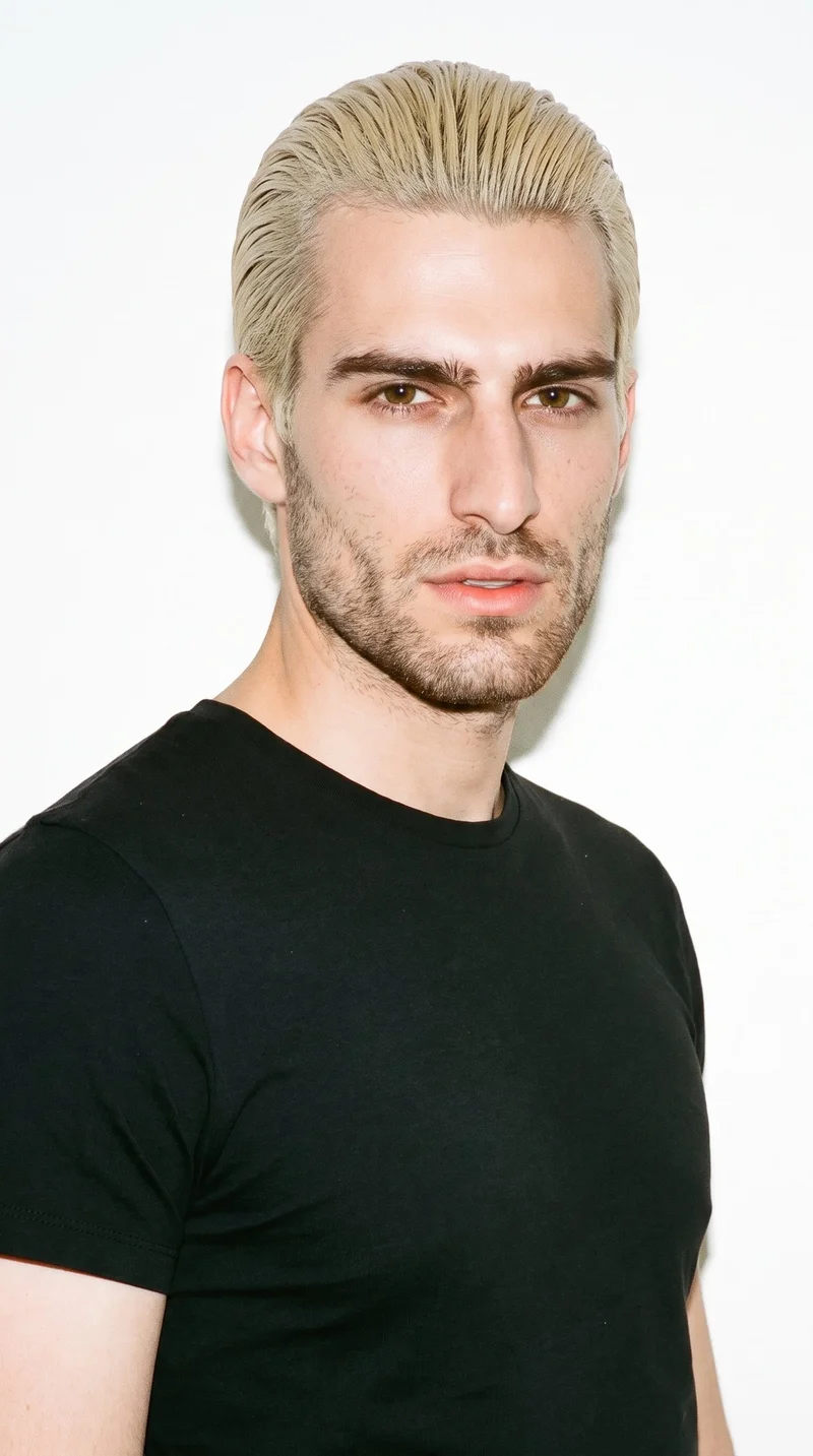 This 23-year-old Caucasian man has fair skin with a neutral undertone and a Roman face shape marked by a prominent nose line and defined jaw. His medium-thickness eyebrows frame brown eyes, and he has thin lips. His very light, near-white blonde hair is combed flat and back close to the head, while dense heavy stubble forms a dark, defined layer across the beard area.
