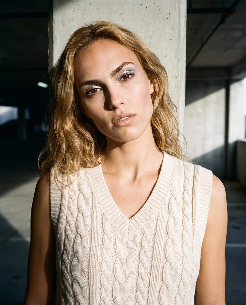 A 19-year-old Caucasian woman wears a cream cable-knit sweater vest in a bust-length front view. Her shoulders are dropped, and she has a curious facial expression. A concrete parking garage serves as the backdrop.