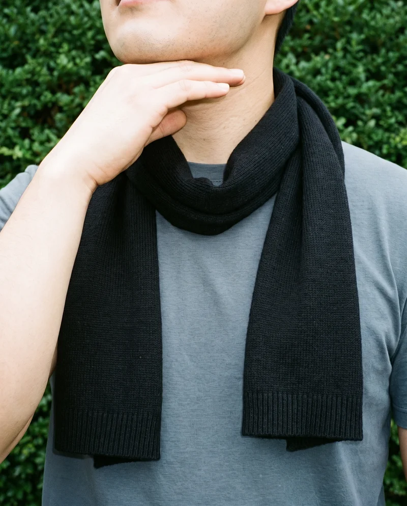 A black wool scarf is wrapped around a man's neck in a front view, with one hand positioned under his jaw. A Zen rock garden serves as the backdrop.