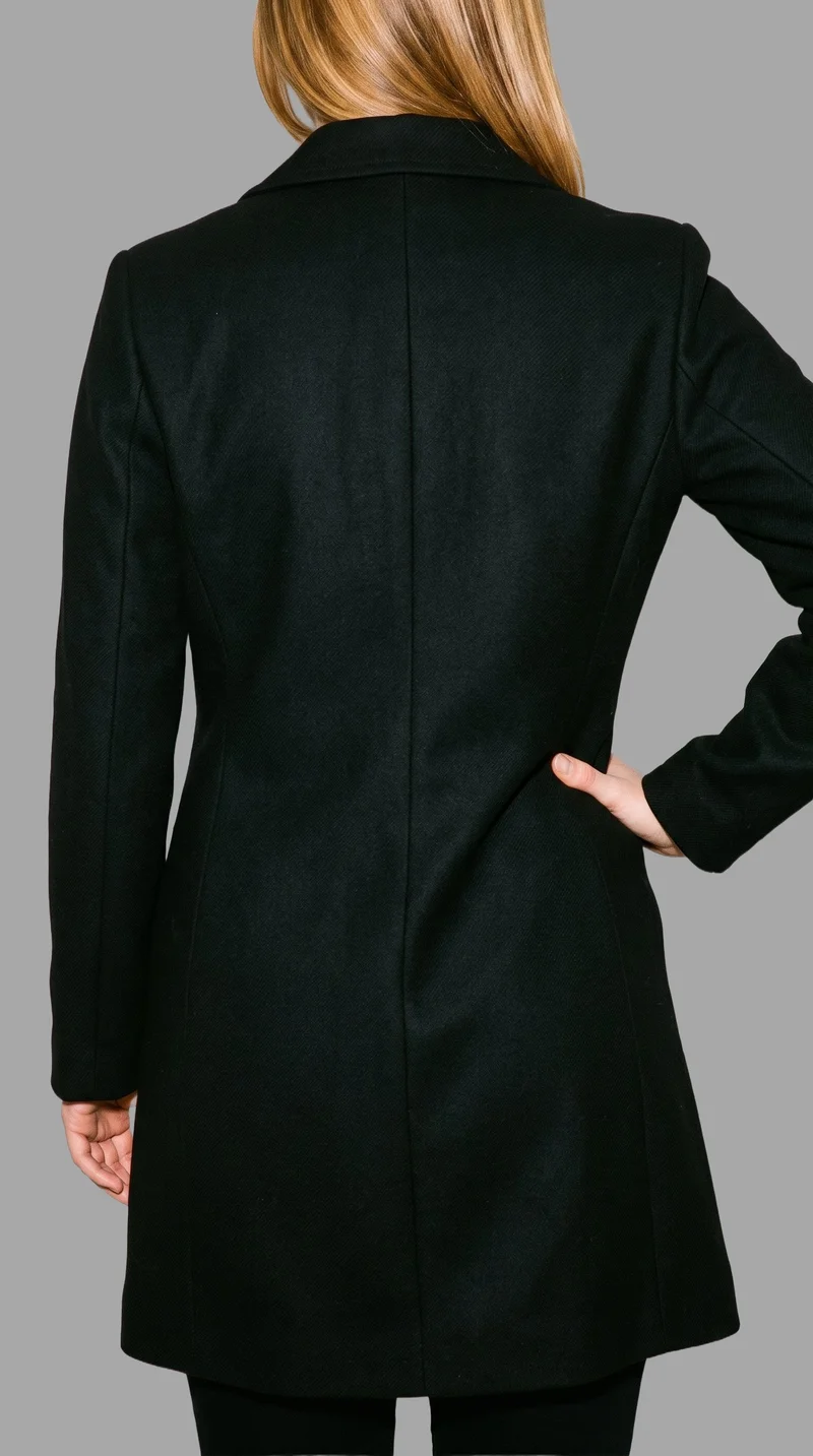 A woman is shown from the rear in a torso-framed view wearing a black overcoat, with one hand resting on her hip. The coat is presented against a flat pebble grey backdrop.