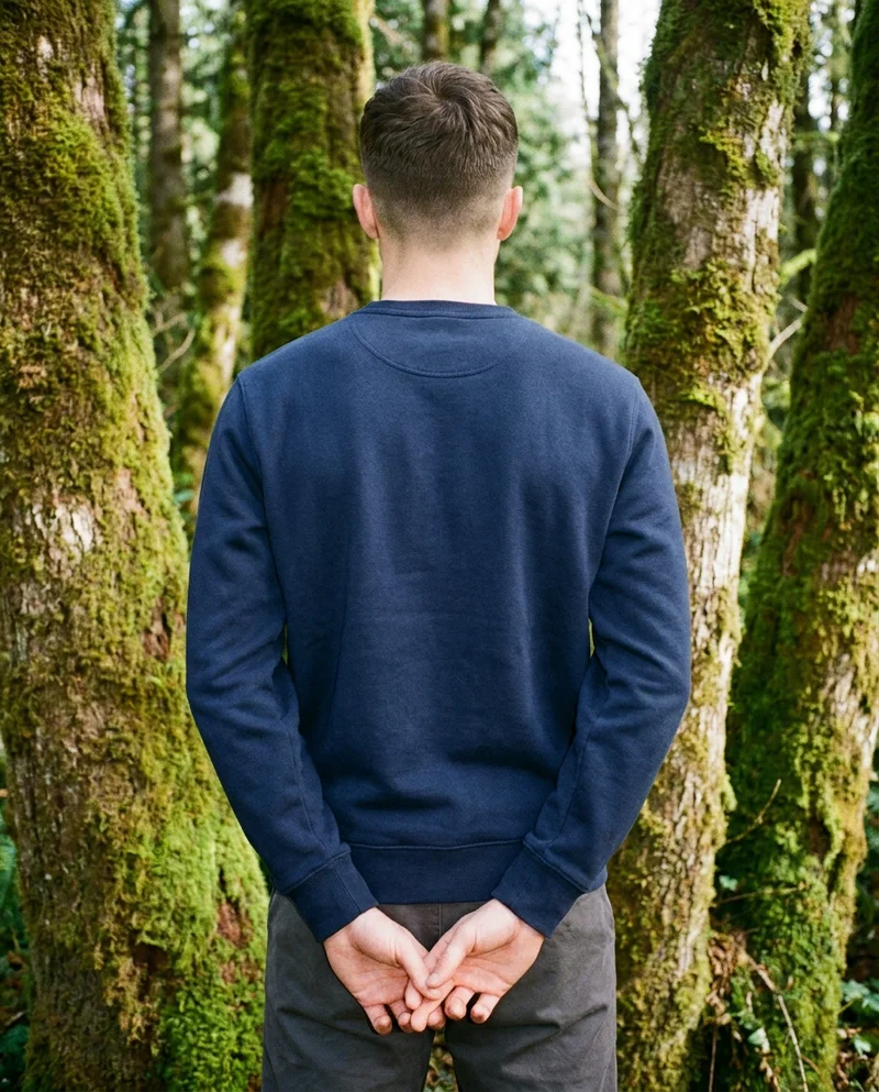 A 31-year-old Caucasian man wears a navy cotton crewneck sweatshirt, shown from the back in an upper-body frame with his hands behind his back. The Olympic Rainforest appears as the backdrop.