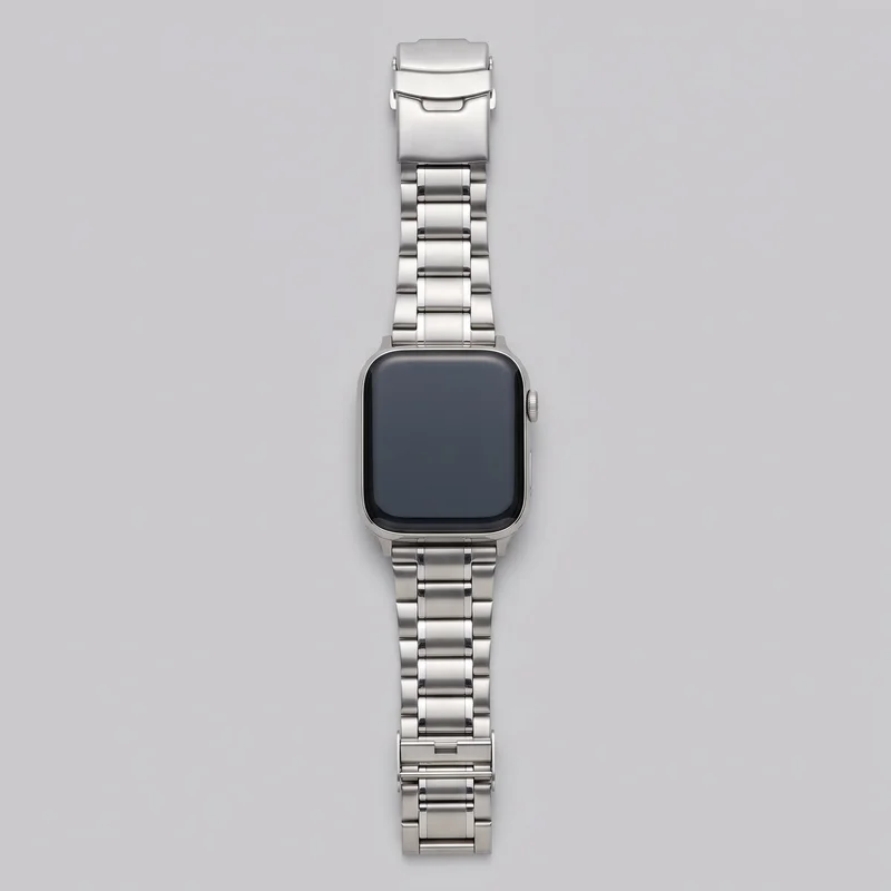 A standalone silver smartwatch lies flat with its articulated multi-link bracelet opened vertically. The rectangular case has rounded corners, a smooth blank display, short integrated lugs, a ridged crown on the right side, and a slim elongated button below it. The bracelet features broad central links with narrower outer links, and the visible fold-over clasp includes a wider hinged cap with a notched release detail and a separate clasp segment.
