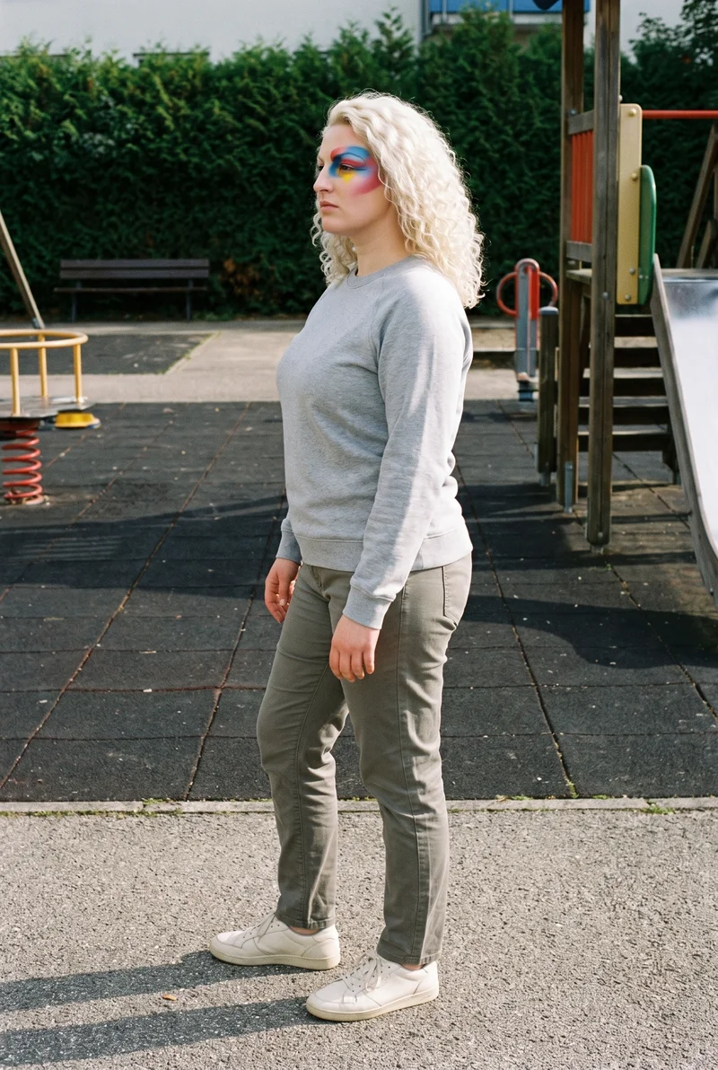 A 32-year-old Caucasian woman wears a grey crewneck sweatshirt in a full-body side view, standing with her weight shifted back and a natural expression. A playground forms the backdrop.