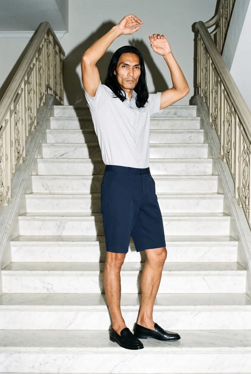 A 40-year-old Southeast Asian man wears navy tailored shorts in a full-body view from a front three-quarter angle. His arms are raised overhead, and he has a confident expression. A marble staircase forms the backdrop.