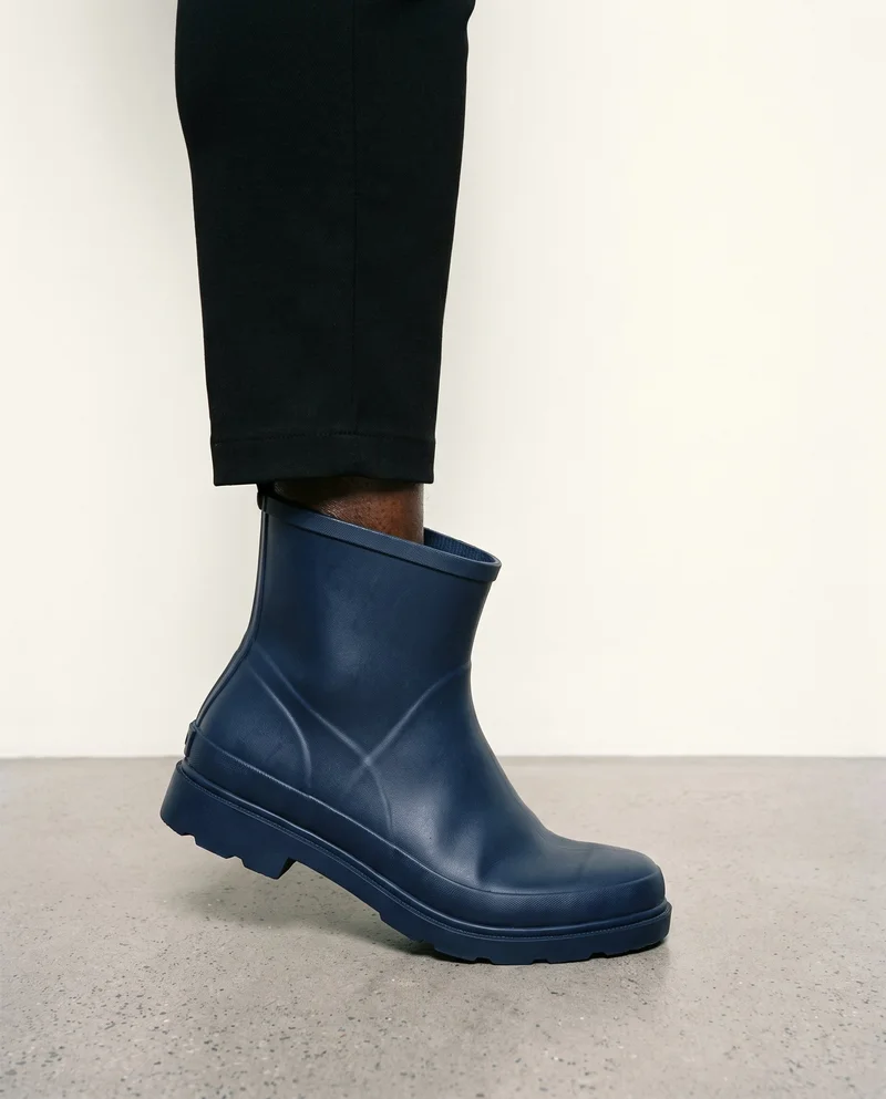 A navy rubber rain boot shown in side view, framed from the ankle while the wearer stands on tiptoes. The boot is set against an alabaster studio-wall backdrop.