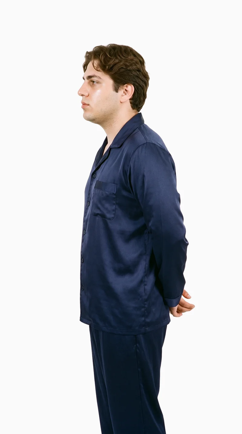 A 20-year-old Turkish man is shown in a three-quarter-length side view wearing a men's satin pyjama set, with his hands behind his back and a confident expression. The image has a flat Seasalt-colored backdrop.