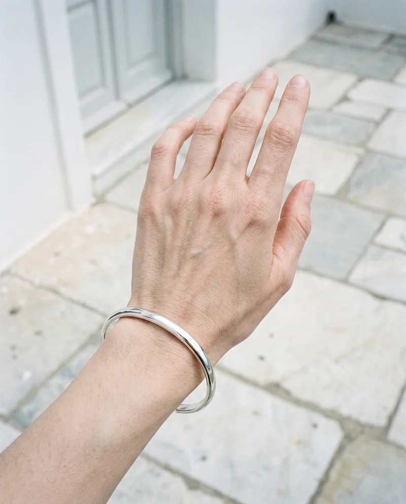A woman's hand and wrist are shown from above at an angle, wearing a silver cuff bracelet. A Santorini caldera view serves as the backdrop.