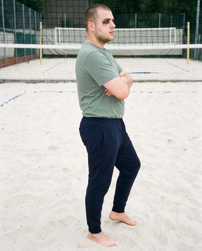 A 25-year-old Slavic man wears navy cotton joggers in a full-body side view, standing in a self-hug pose with a neutral expression. A beach volleyball court forms the backdrop.