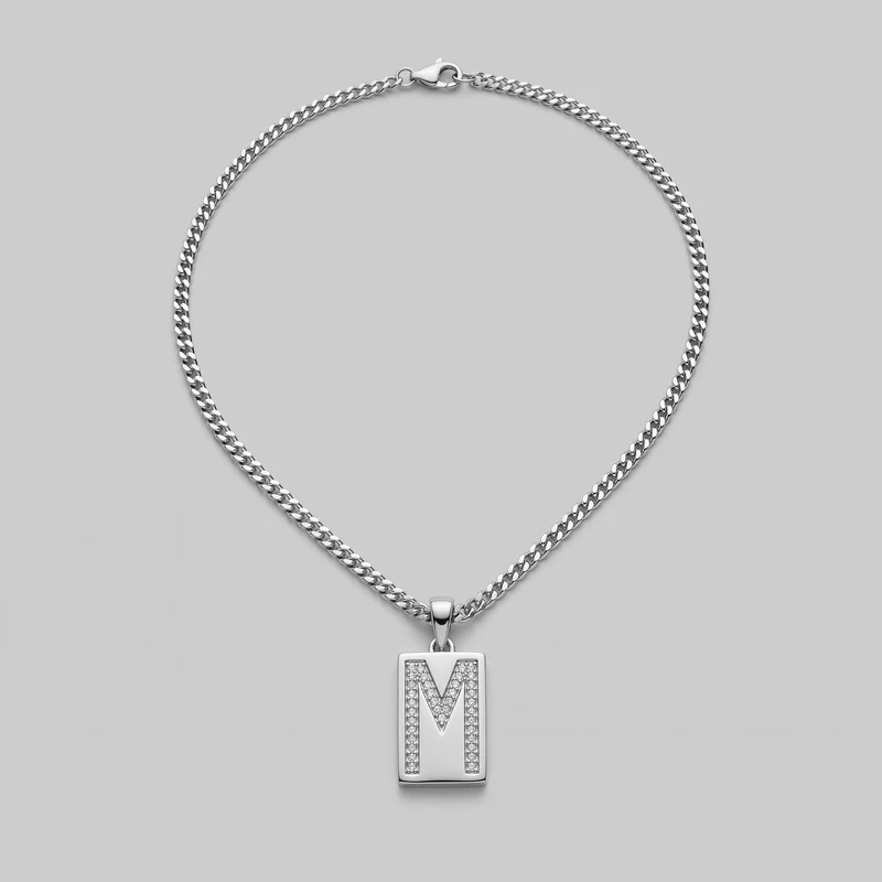 A standalone curb-link pendant necklace is arranged in a rounded loop, with a rectangular pendant centered at the bottom. The pendant has a beveled raised border and a block letter “M” filled with pavé-set stones. A lobster-claw clasp and jump rings are visible at the top, and a smooth bail connects the pendant to the chain.