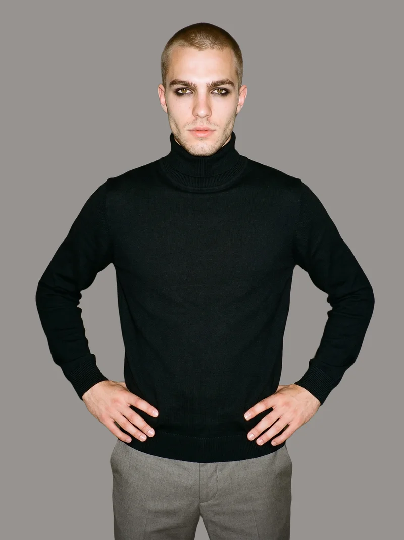 A 20-year-old Caucasian man wears a black turtleneck in a three-quarter-length frame. He is shown from a front three-quarter angle with his hands on his waist and a natural facial expression against a flat grey studio backdrop.