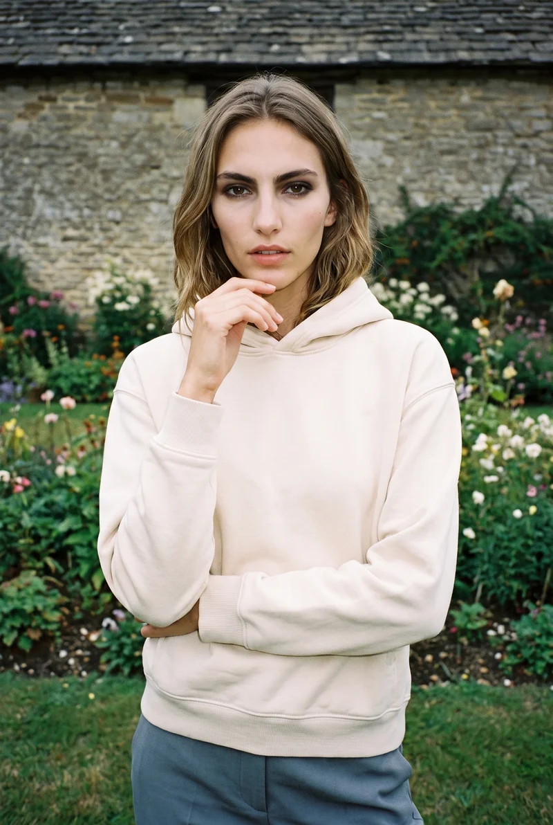 A 19-year-old Caucasian woman is shown from the front in an upper-body view wearing a cream pullover hoodie. She holds one hand at her chin and has a serious expression. A cottage garden forms the backdrop.