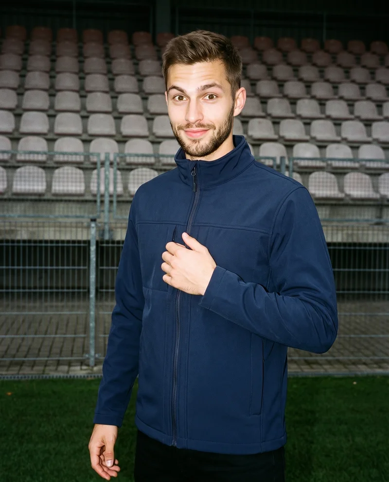A 22-year-old Caucasian man wears a navy softshell jacket in an upper-body view from a front three-quarter angle, with his fingers at his chest and a curious expression. Stadium seating forms the backdrop.