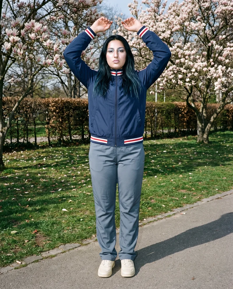 A 19-year-old Turkish woman wears a women's navy retro track jacket in a full-body front view, standing with her arms overhead and a neutral expression. A Magnolia Tree Park backdrop is visible.
