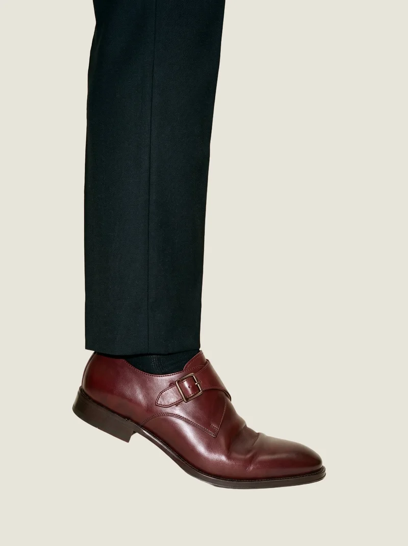 Oxblood monk-strap shoes shown in an ankle-framed side view while positioned on tiptoes, against an oyster pearl flat-color backdrop.