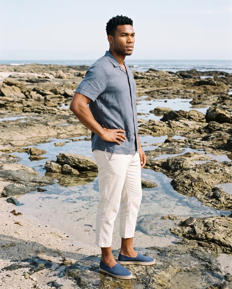 A 28-year-old African man is shown full body in side view wearing navy canvas espadrilles, with one hand on his hip and a natural expression. A rocky beach forms the backdrop.