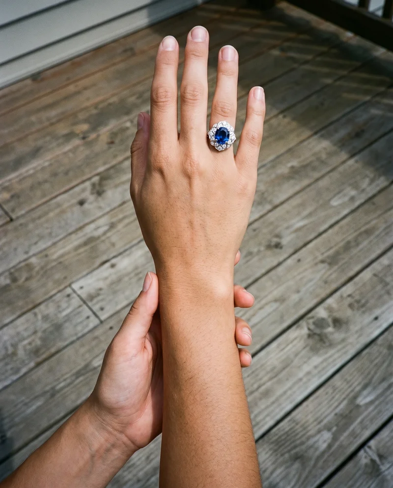 A hand and wrist are shown from a top view while the hand touches the wrist, displaying a gemstone statement ring. A backyard deck serves as the backdrop.