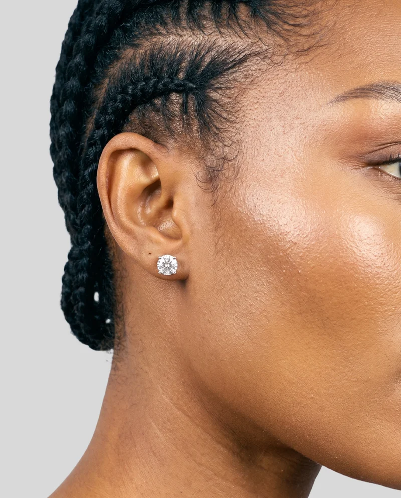 A woman's ear is shown in side view, angled toward the shoulder, wearing a diamond stud earring. The image uses a pearl grey flat-color backdrop.