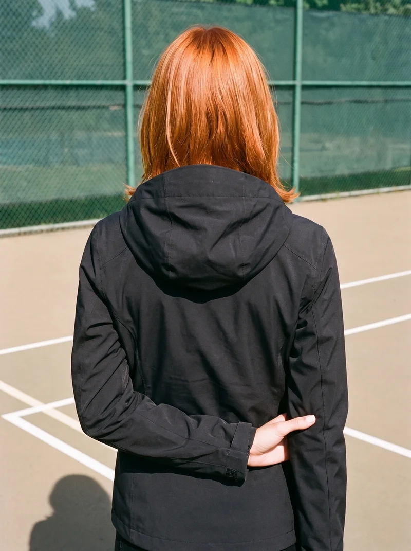 A 21-year-old Caucasian woman wears a black women's hardshell jacket, shown from the back in an upper-body frame while touching her wrist. A tennis court serves as the backdrop.