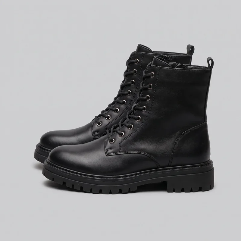 A pair of women's black combat boots is shown in a clear side three-quarter view. The boots have high ankle shafts, rounded closed toes, plain vamps, front lace closures with multiple round eyelets, central tongues, visible side zippers near the shaft openings, and rear pull loops. Stitched panel seams, raised welt edges with visible stitching, low block heels, and thick deep-tread lugged outsoles are visible.