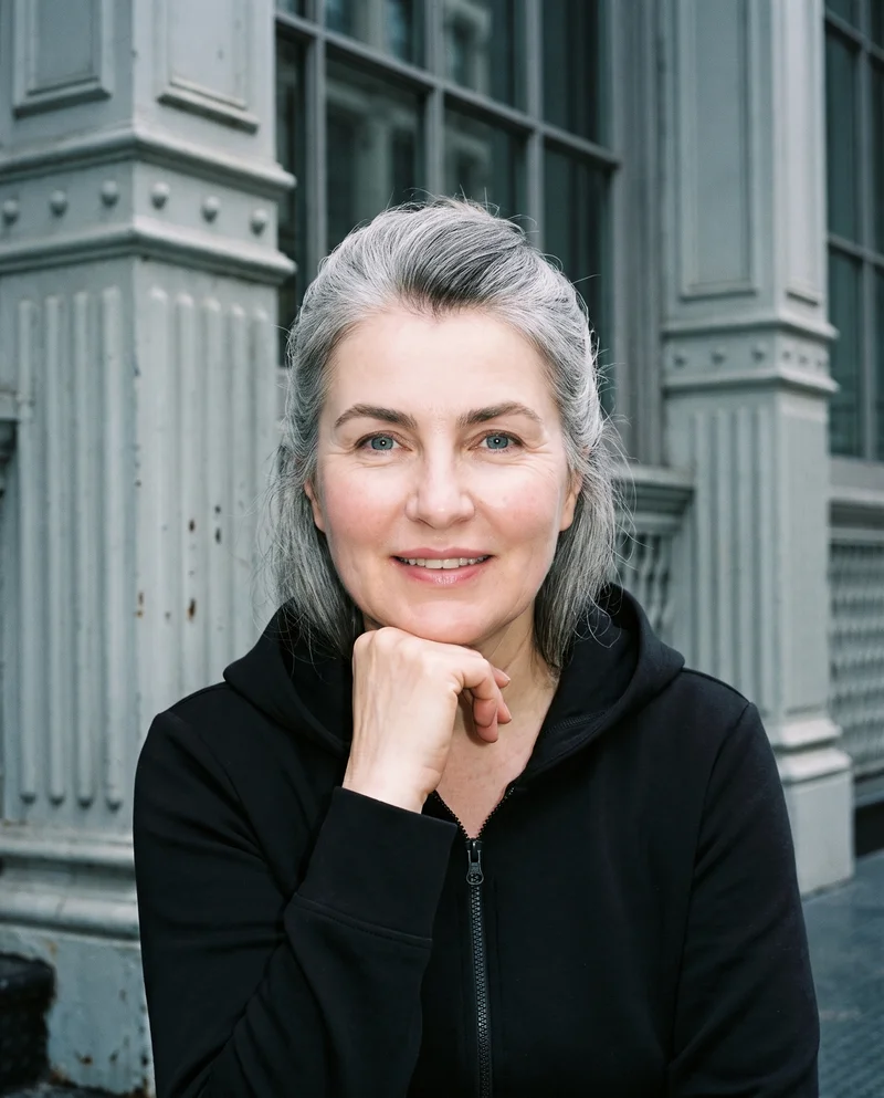 A 54-year-old Slavic woman wears a black zip-up hoodie in a bust-length front view. She smiles with her chin resting on her knuckles, against a Cast Iron District cobblestone backdrop.