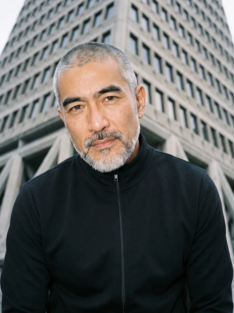 A 50-year-old Japanese man wears a black tracksuit top in a bust-length frame, shown from a front three-quarter angle with a shoulder-drop pose and a neutral expression. A pyramid building appears as the low-angle backdrop.