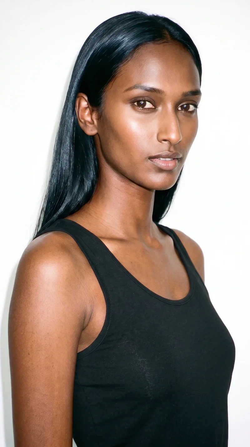 An 18-year-old Indian woman with dark-brown skin and a warm golden undertone. Her face is long, with more length than width, a tall forehead, and elongated cheeks. She has thick eyebrows, brown eyes, and medium-sized lips. Her deep black hair is long and falls straight without waves or layers.