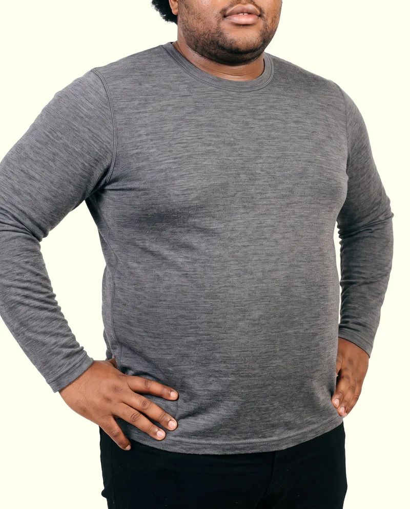 Men's grey merino thermal top shown on a male model in an angled front torso view, with his hands on his hips. An ivory lace backdrop frames the studio-cutout photograph.
