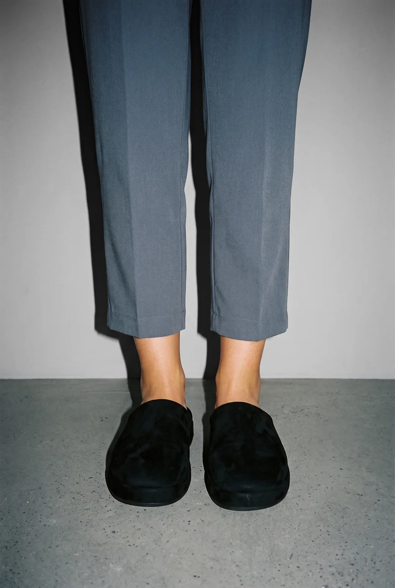 A woman's lower body is shown from the front with her feet together, wearing black memory-foam slippers. A pearl grey studio-wall backdrop fills the background.