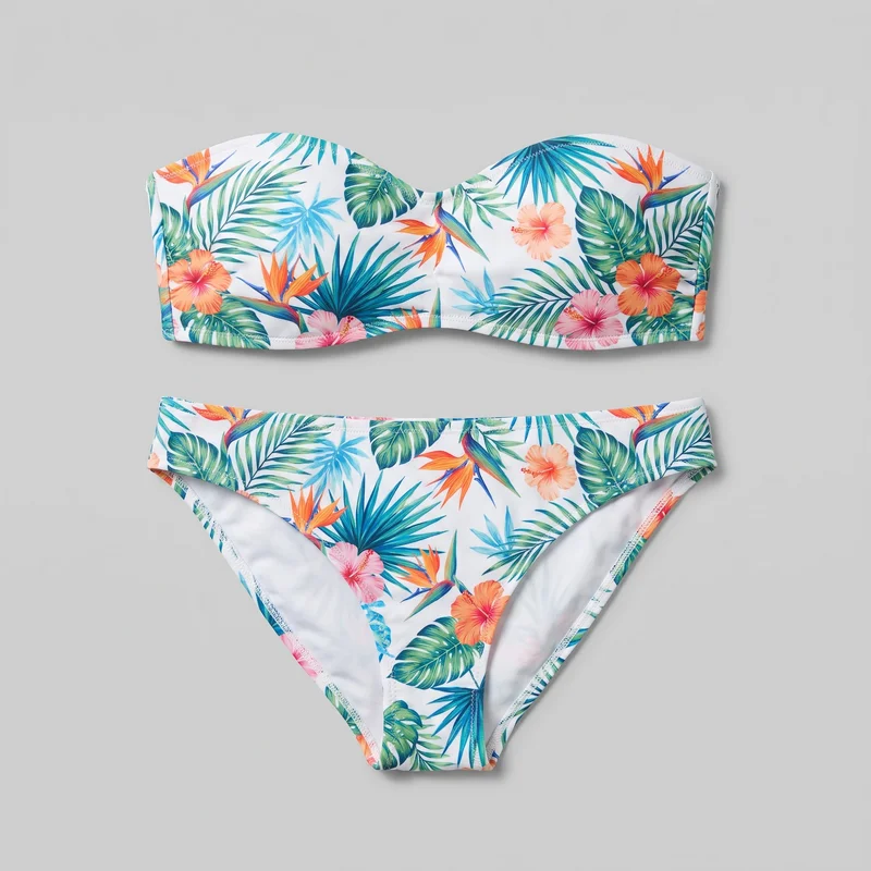 A complete matching two-piece bikini set is arranged in a flat lay, with a strapless bandeau top positioned above a bikini brief bottom. The front-facing view shows the shaped top and high-cut brief outline, both covered in a tropical botanical print featuring palm and monstera-style leaves, hibiscus flowers, and bird-of-paradise motifs.