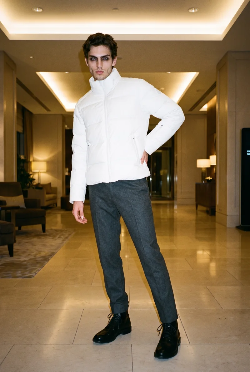 An 18-year-old Caucasian man wears a white padded ski jacket in a full-body front three-quarter view. One hand rests on his lower back, and his natural facial expression is visible against a hotel lobby backdrop.
