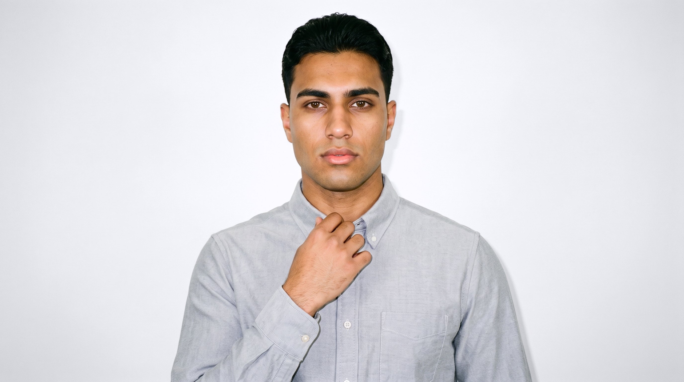 A 19-year-old Arab man is shown from the front in a bust framing, wearing a grey cotton Oxford shirt with one hand at his collarbone. He has a neutral facial expression, set against a porcelain studio-wall backdrop.