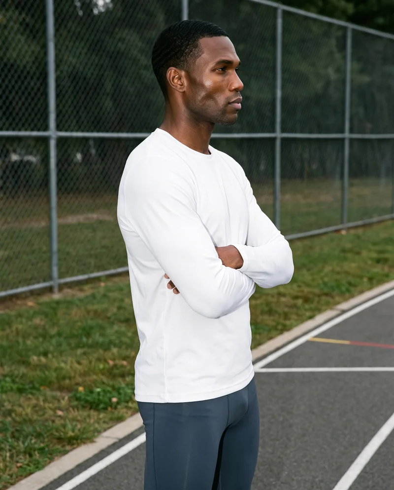 A 42-year-old African man wears a men's white performance base layer in a three-quarter-length side view, standing with his arms crossed and a natural expression. A running track serves as the backdrop.