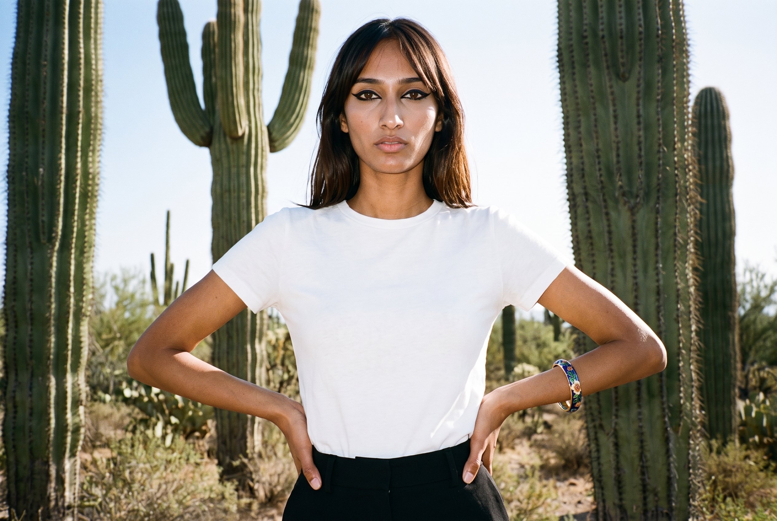 A 20-year-old Indian woman is shown from the front in an upper-body view, wearing an enamel bangle with her hands on her hips and a neutral expression. A cactus desert backdrop is visible behind her.