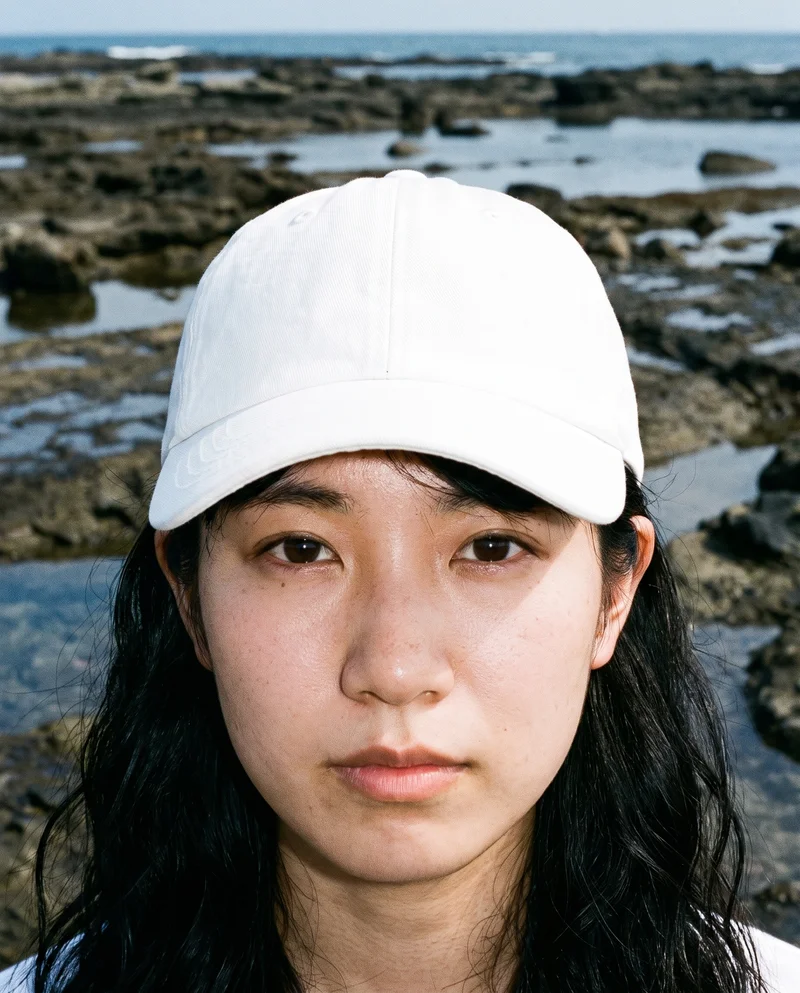 A 22-year-old Japanese woman wears a white baseball cap, shown from the front with her head level and a natural expression. The head-only composition has a Tidal Pool Rocks backdrop.